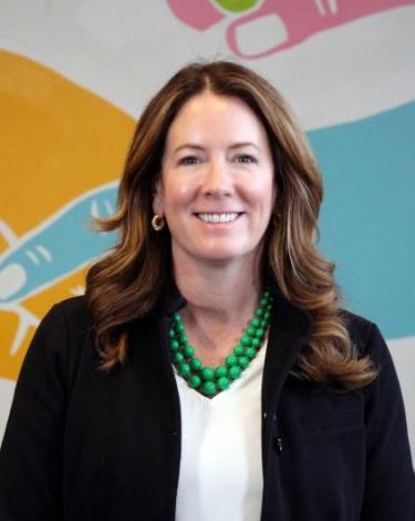 Kathy smiling in black blazer, white shirt, green pears against colorful wall