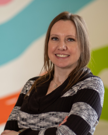 Alicia smiling in black and gray stripe shirt against colorful wall