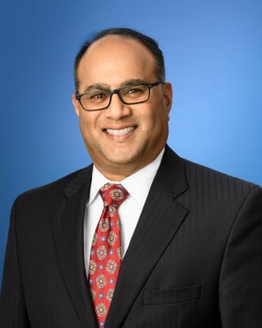 Alan in black blazer with red tie against blue background